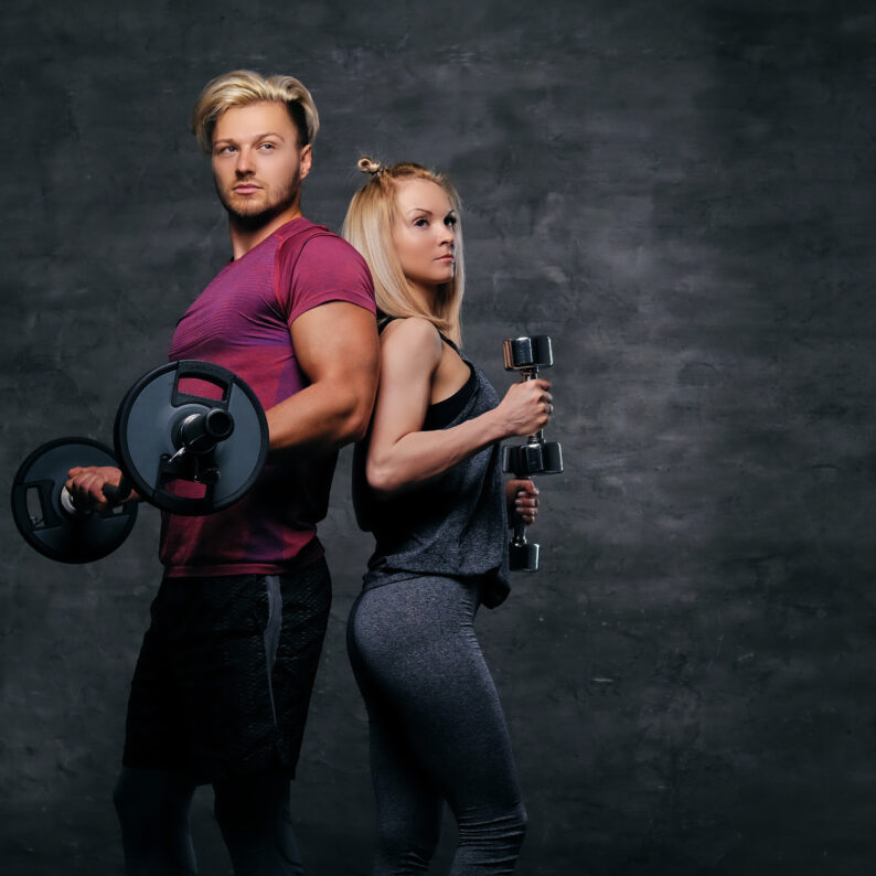 An attractive fitness couple of sporty male holds barbell and slim blond female holds dumbbells over grey background